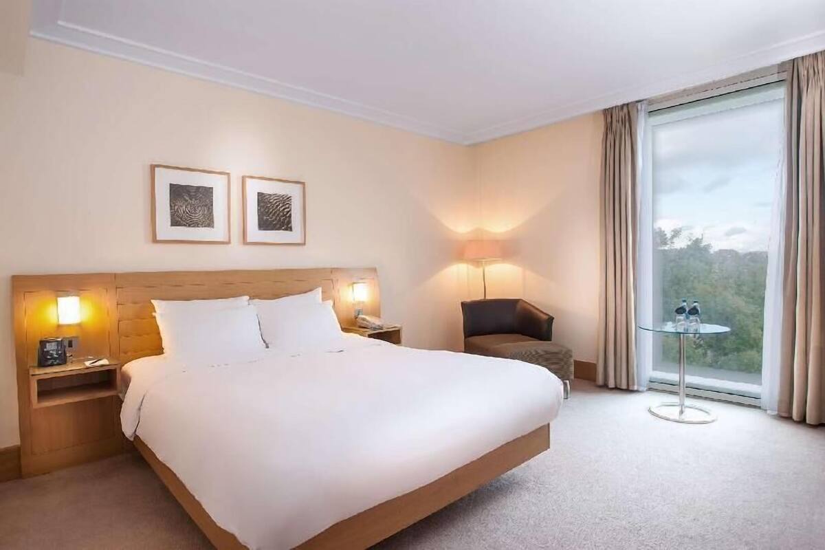 Hotellihuone, jossa on suuri sänky, yöpöytälamput, tuoli, pieni pöytä pulloineen ja seinällä tauluja.