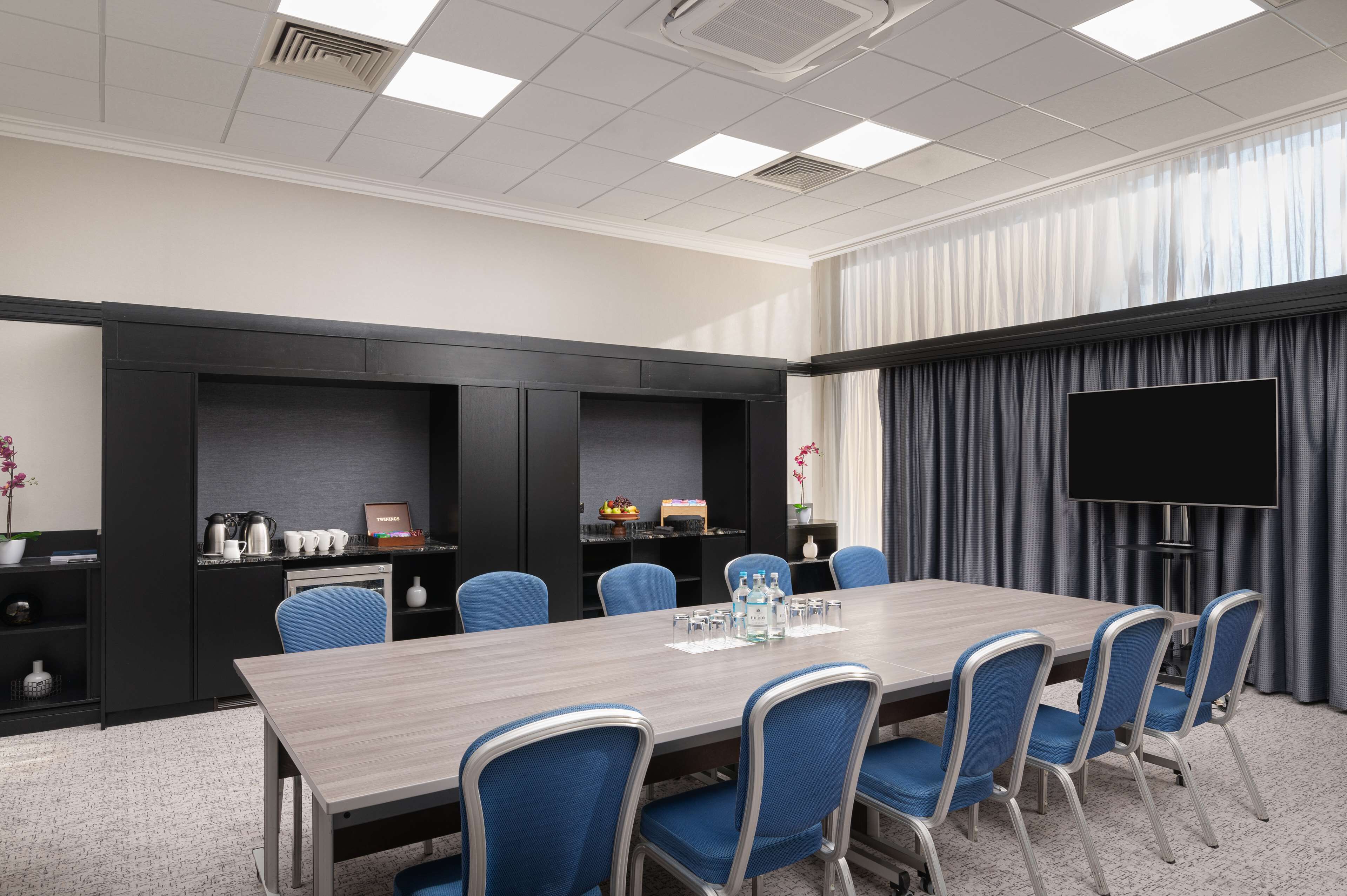 A conference room with a long table, blue chairs, a flat-screen TV, and a large window with curtains.