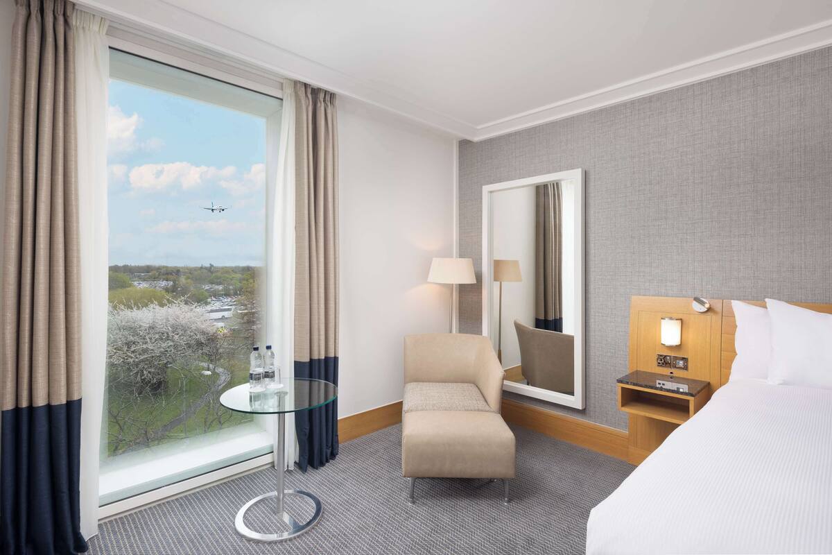 Hotellihuone, jossa on suuri ikkuna, sänky, tuoli, pieni pöytä, yöpöytä ja peili.
