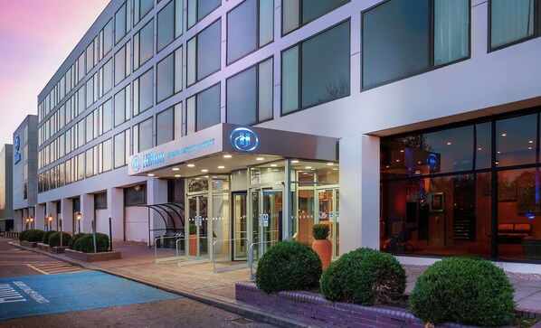 A modern building with a glass facade, a sign for Hilton London Gatwick Airport, and a blue parking bay. - Hilton London Gatwick Airport (Gatwick)