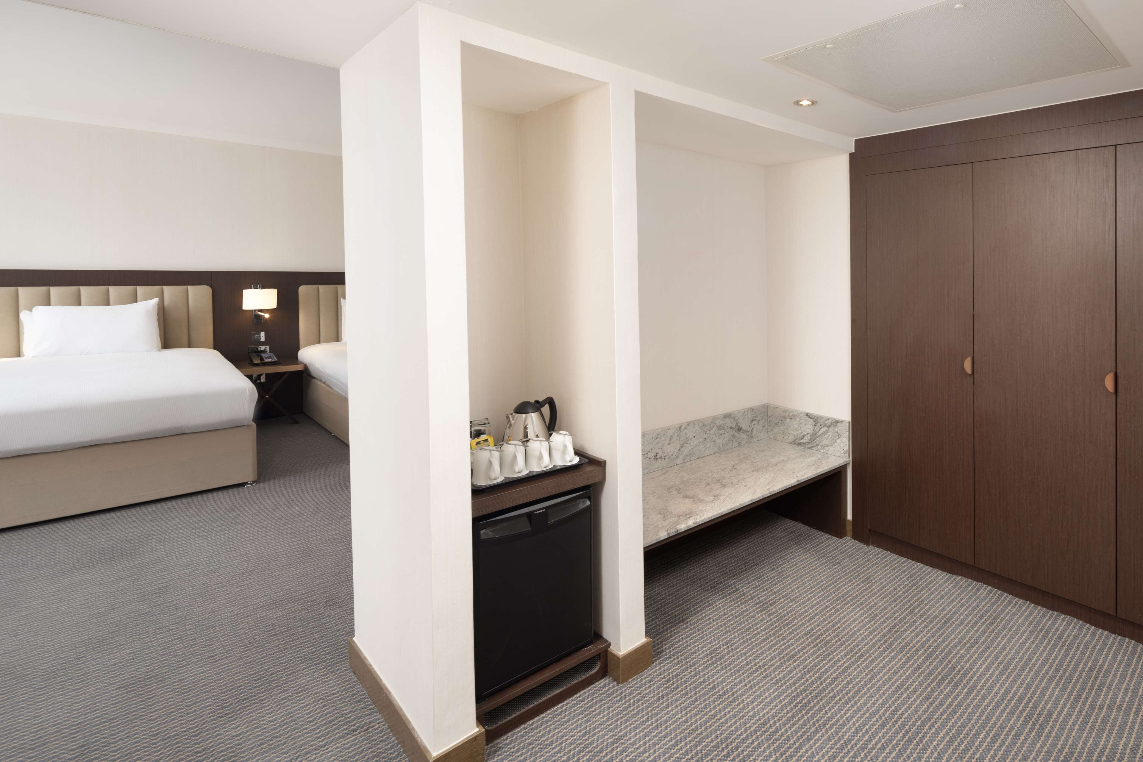 Ein Hotelzimmer mit einem Bett, einem kleinen Schreibtisch mit Lampe, einer Kaffeestation, einer Bank und einem Kleiderschrank.