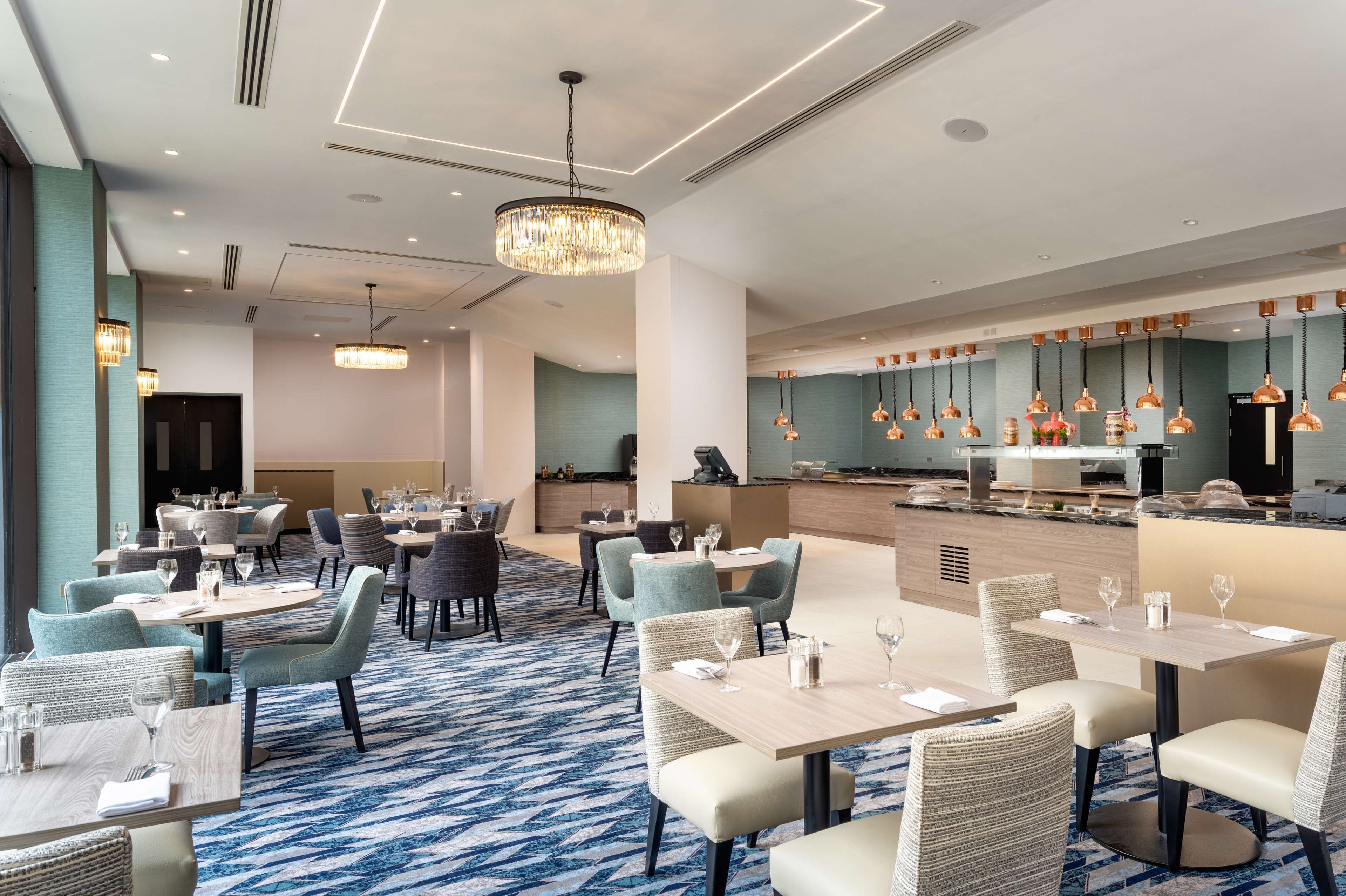 A modern hotel dining area with multiple tables and chairs, a central bar area, and a patterned carpet.