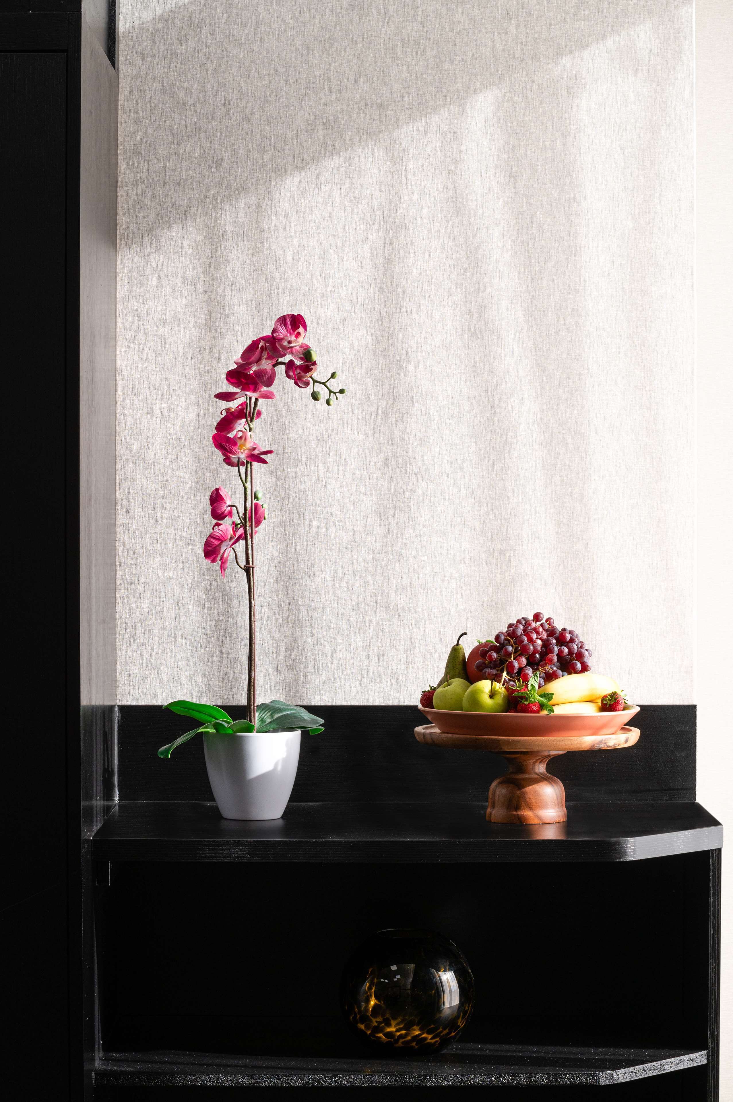 Une orchidée en pot, un panier de fruits et une sphère de verre décorative, le tout posé sur une étagère noire devant un mur blanc.