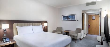 A hotel room with a large bed, two armchairs, a small table, a desk with a telephone, a TV, and a framed picture on the wall.