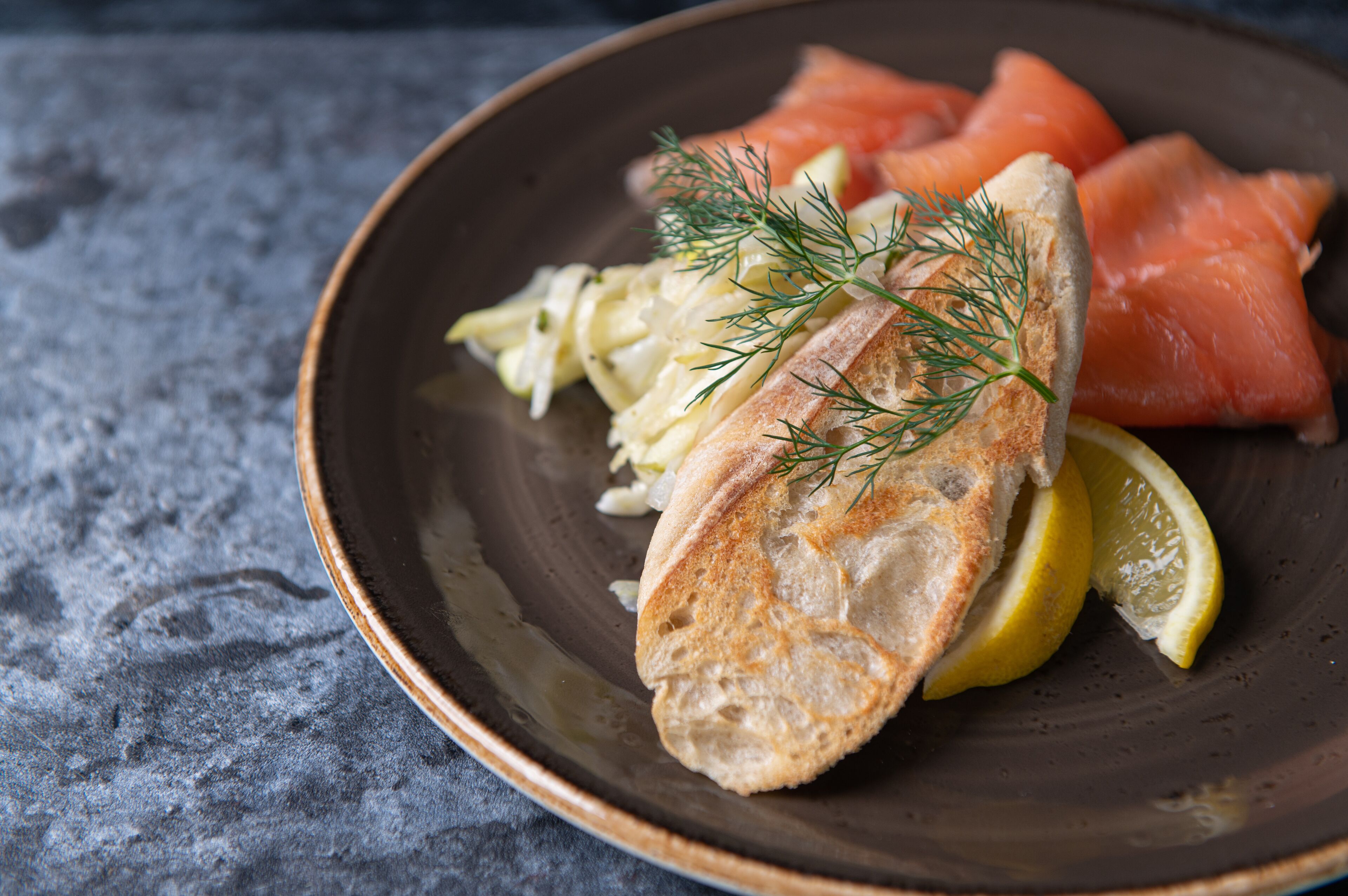 Un plato con salmón ahumado, una rebanada de pan, un gajo de limón y una ramita de eneldo.