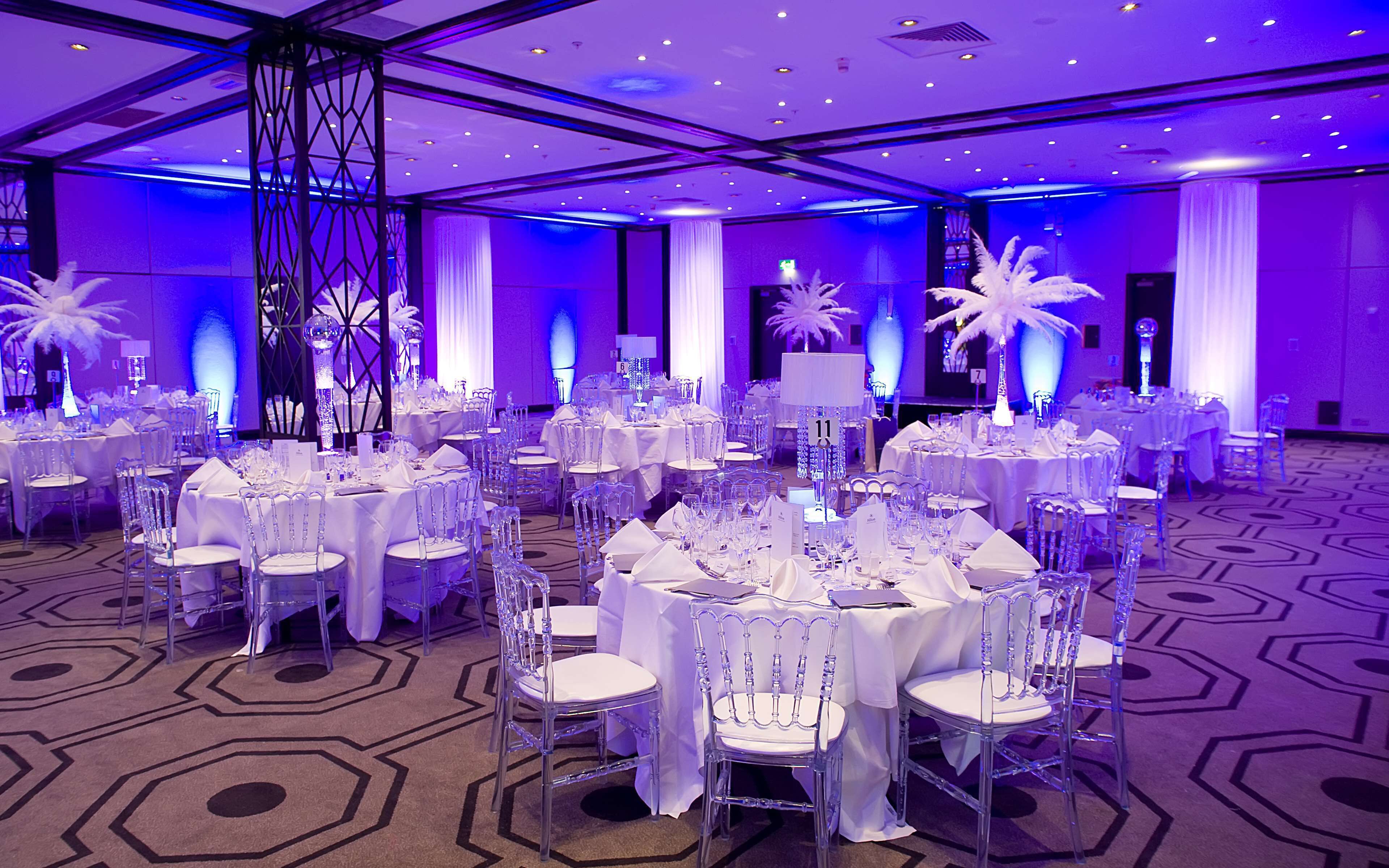 A large banquet hall with round tables, clear chairs, and white tablecloths.