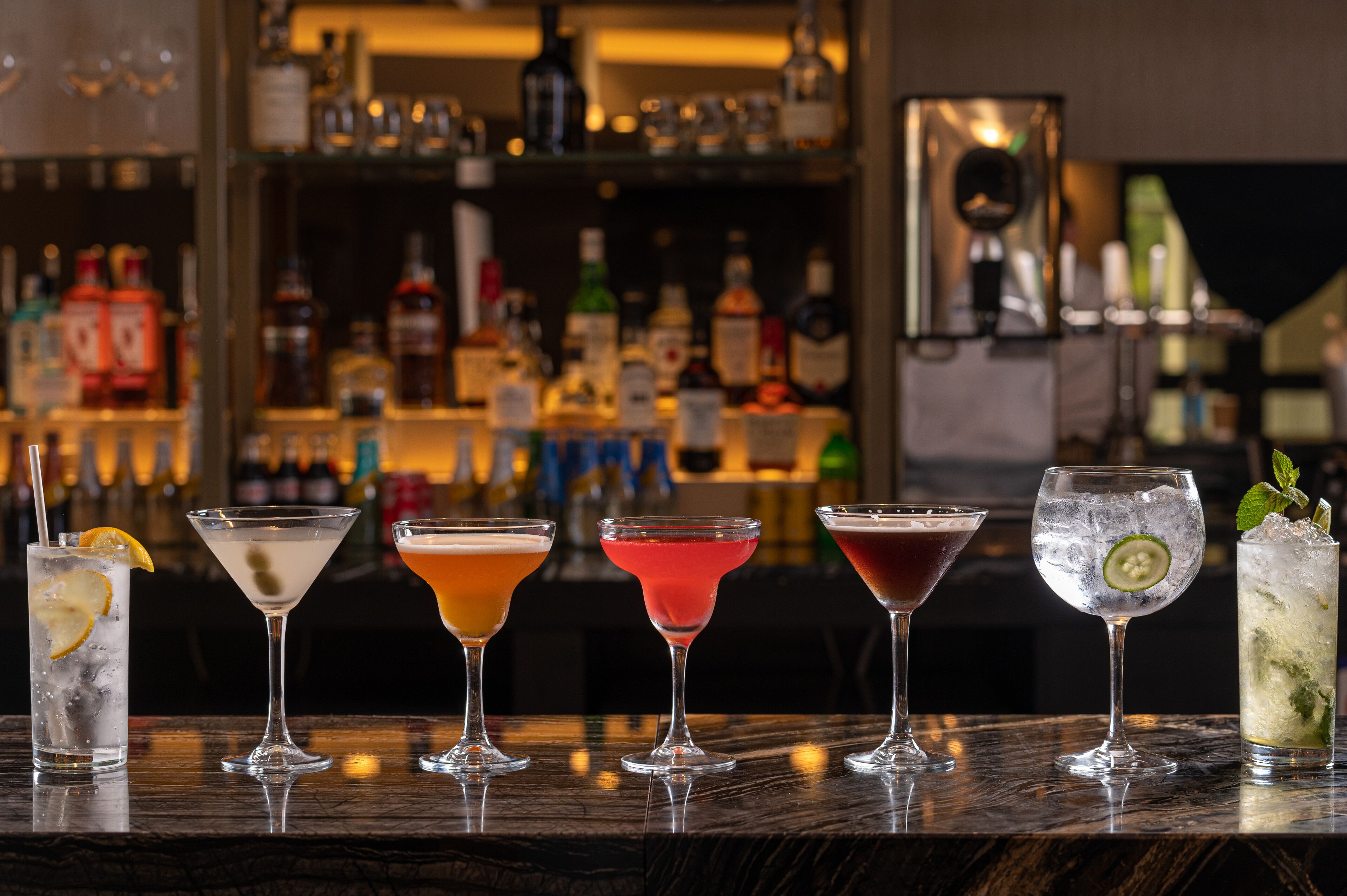 Um bar com diversos coquetéis em taças de martini, uma seleção de garrafas ao fundo e um espelho refletindo a área do bar.