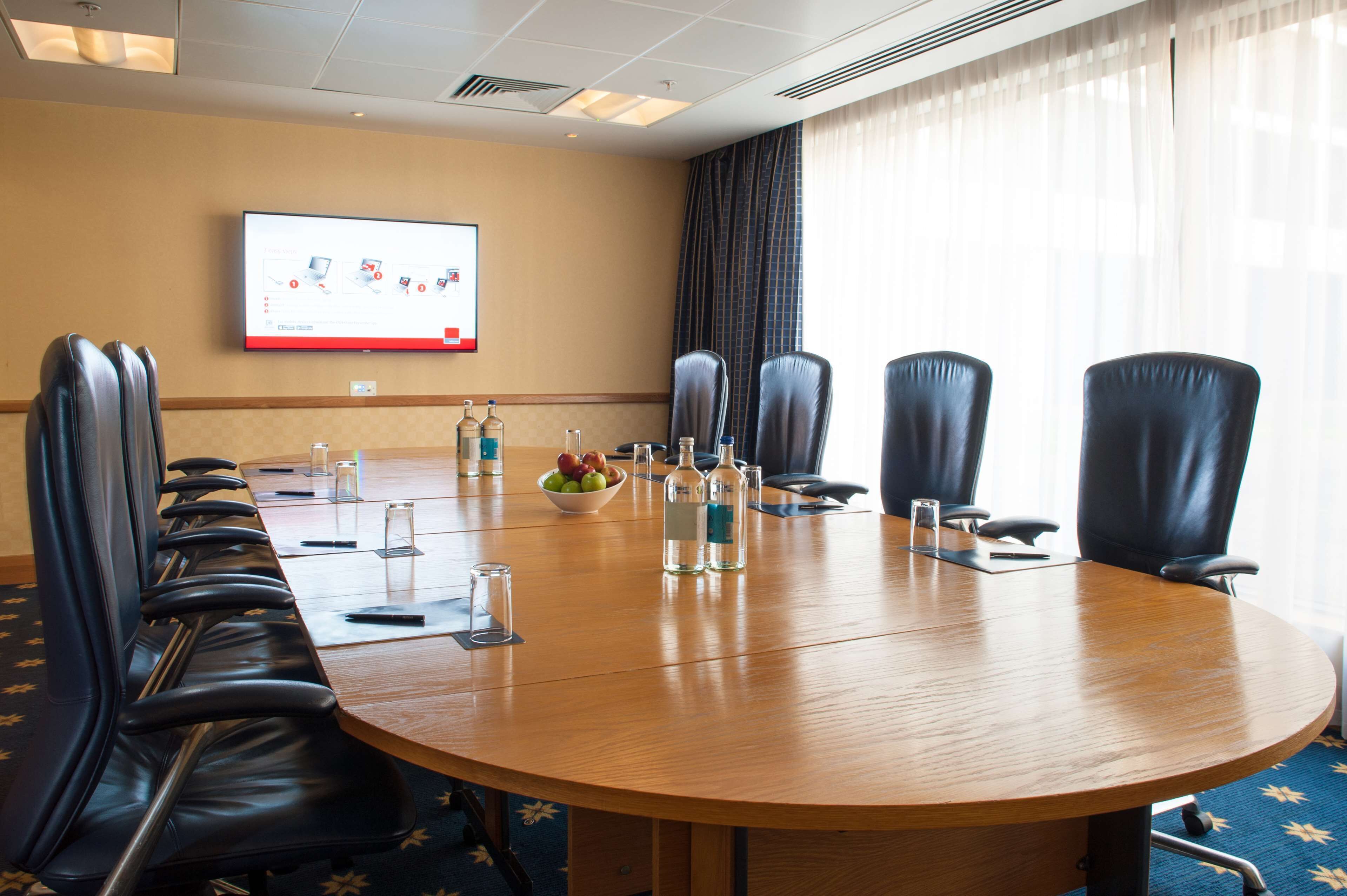 A conference room with a long table, black leather chairs, a projector screen, and a bowl of fruit.