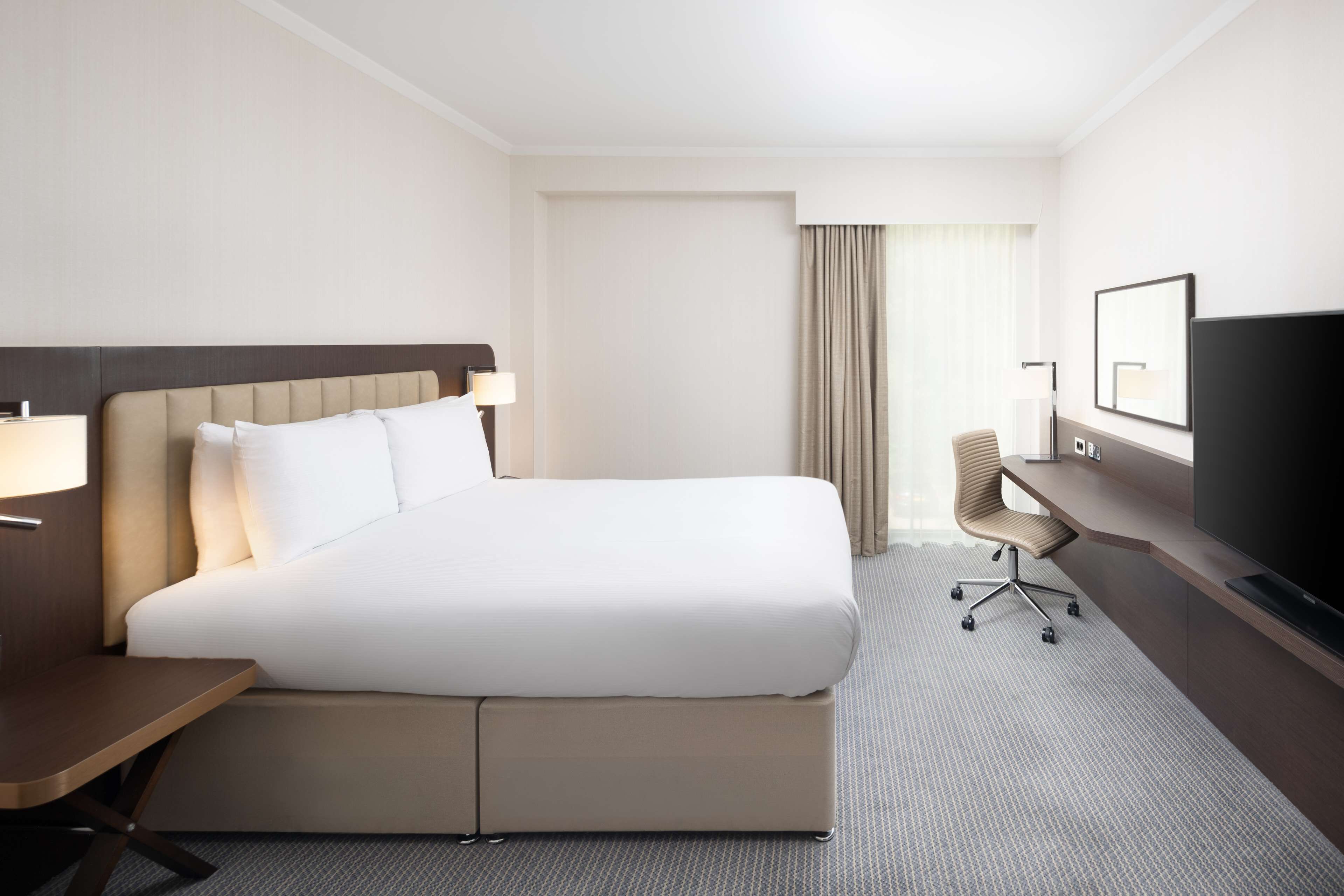 Une chambre d’hôtel avec un grand lit, un bureau avec une chaise, une télévision et une fenêtre avec des rideaux.