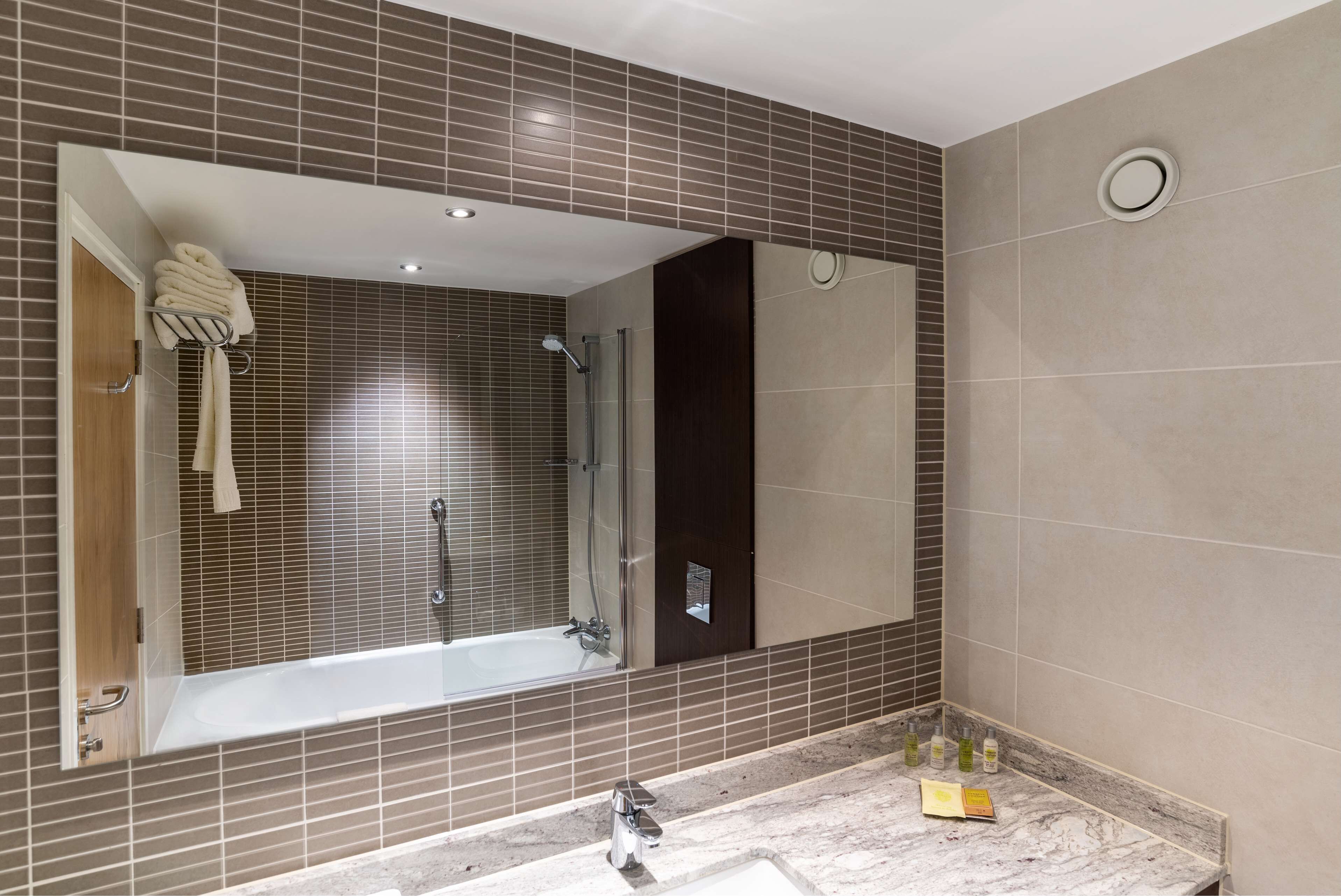 A modern bathroom with a large mirror, a bathtub, a shower, and a marble countertop with toiletries.