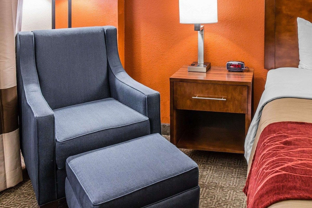 Een hotelkamer met een enkel bed, een nachtkastje met een lamp, een stoel en een klein tafeltje met een klok.