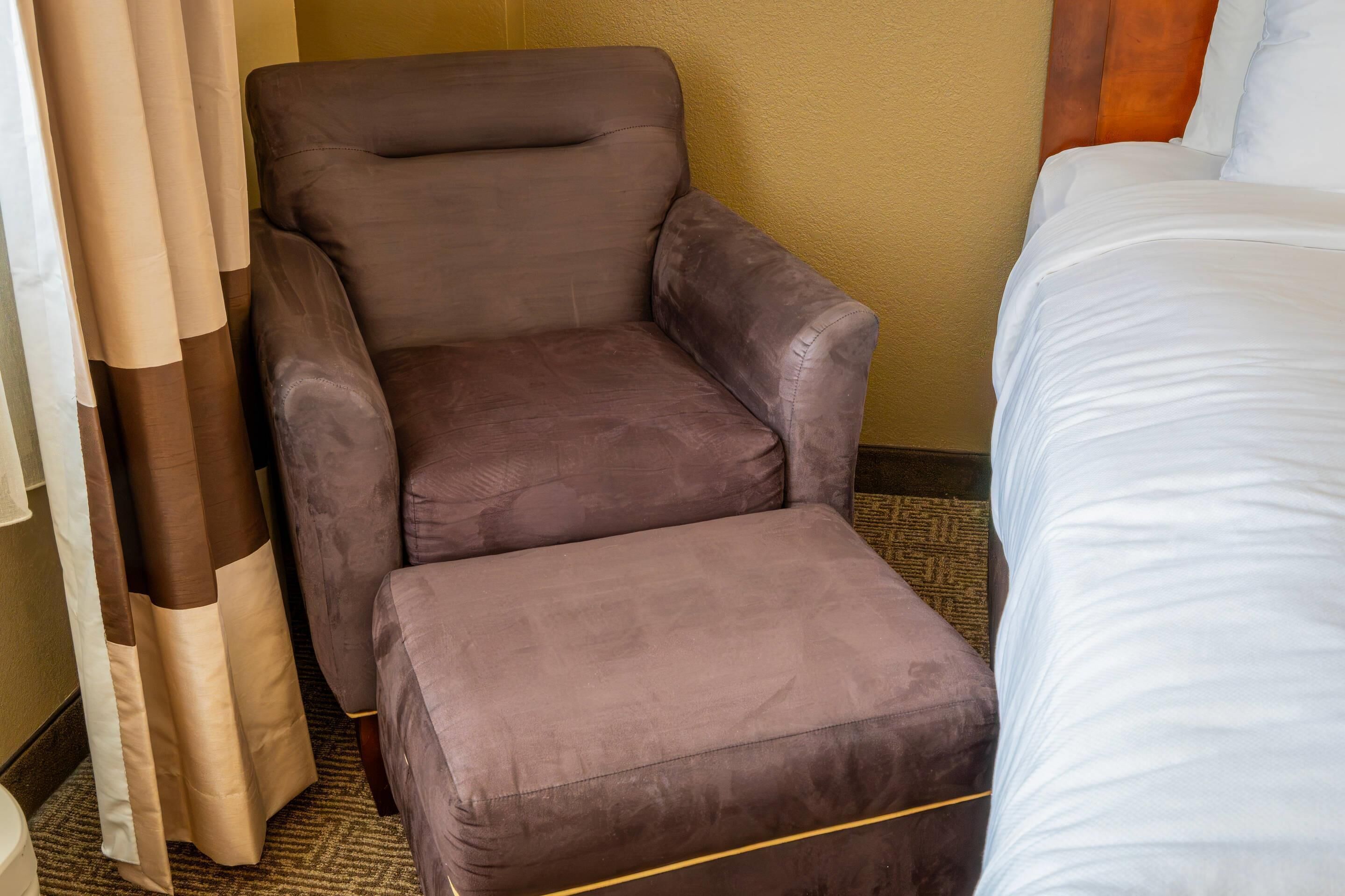 Habitación de hotel con un sillón marrón, un reposapiés a juego, una cama con ropa blanca y una cortina parcialmente abierta.