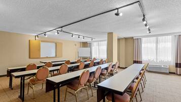 Un salón de conferencias con mesas largas, sillas, una pizarra y una pantalla de proyector.