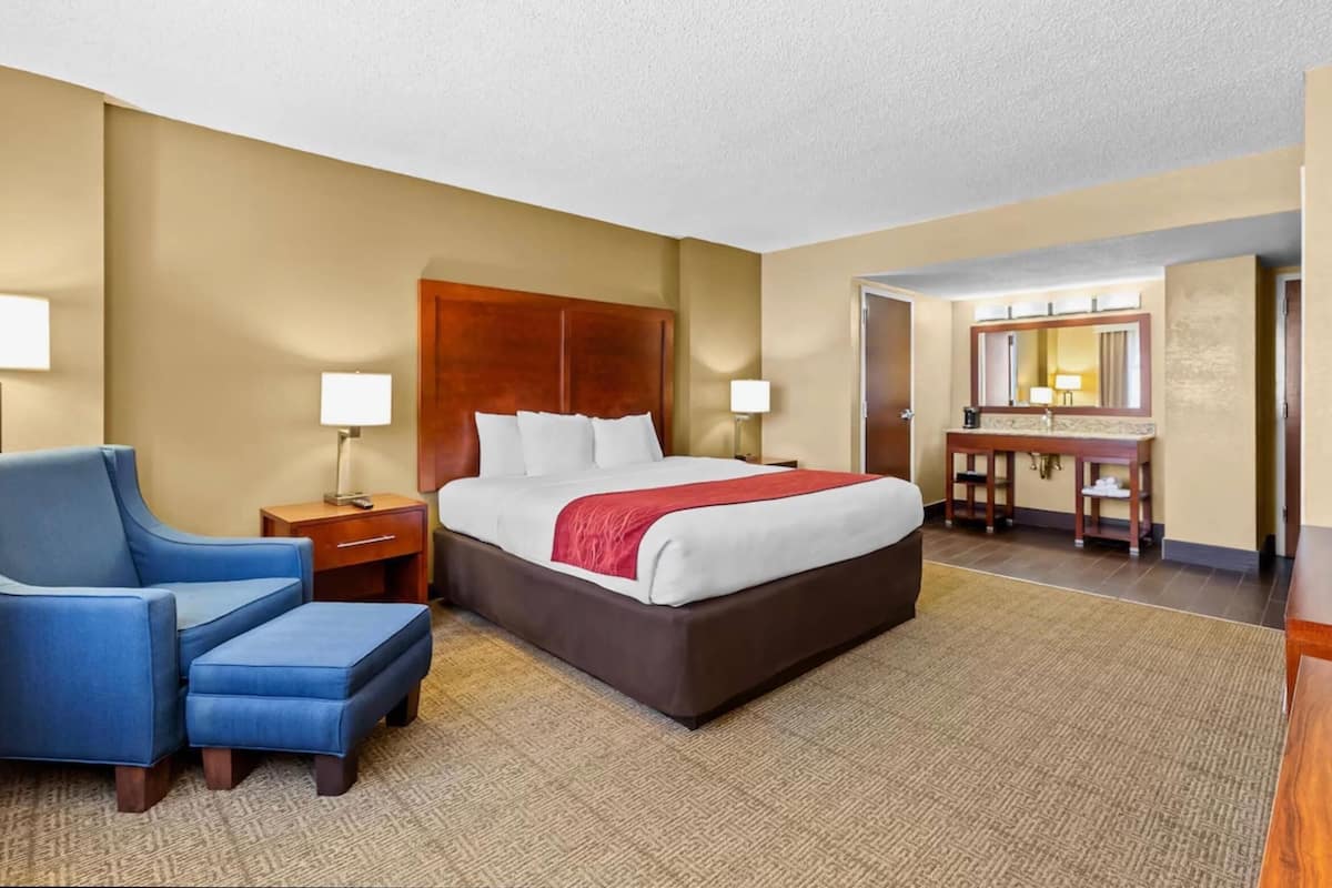 Hotelkamer met een groot bed, een blauwe fauteuil, een nachtkastje, een bureau met een spiegel, en een badkamer zichtbaar door een geopende deur.