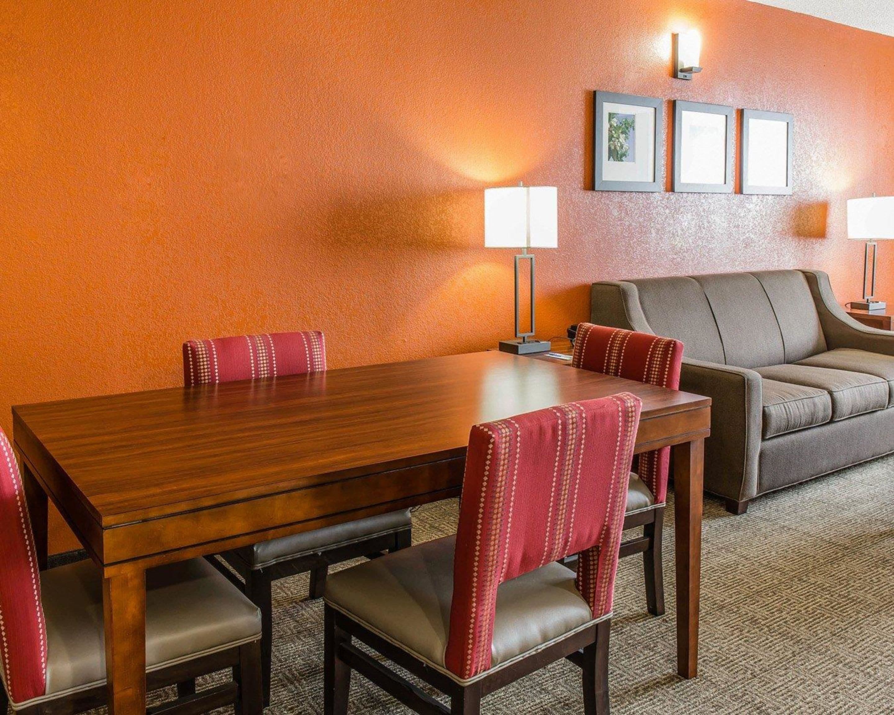 A hotel room with a wooden dining table, four upholstered chairs, a sofa, and two wall-mounted lamps.