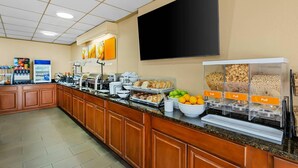 A hotel breakfast area with a long counter, self-service food stations, and a large flat-screen TV mounted on the wall.