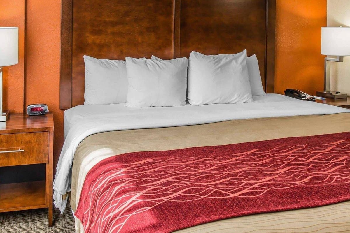 Een hotelkamer met een groot bed, een houten hoofdbord, een rode geborduurde plaid en een nachtkastje met een lamp en telefoon.