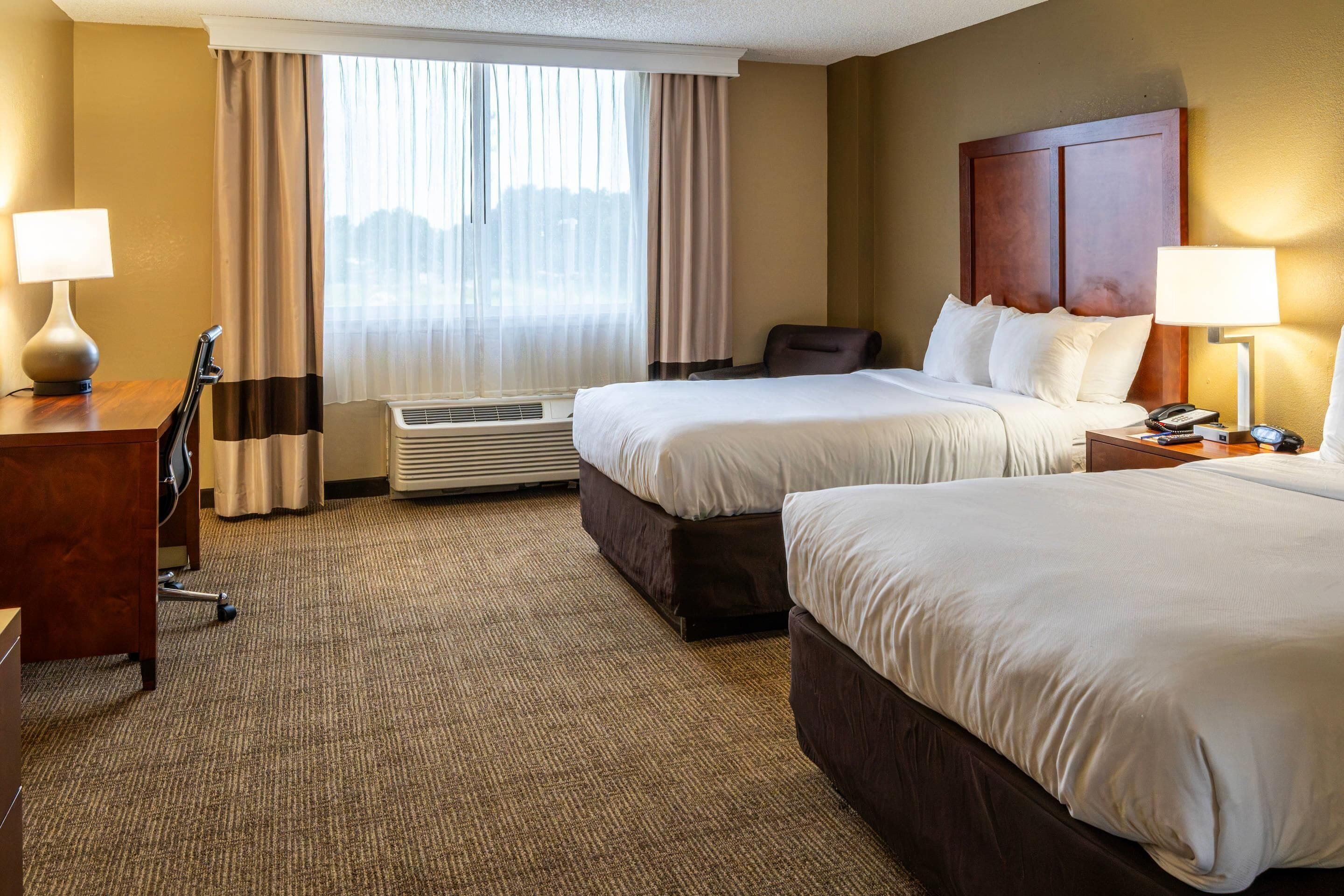 A hotel room with two beds, a desk, a chair, a lamp, and an air conditioning unit.