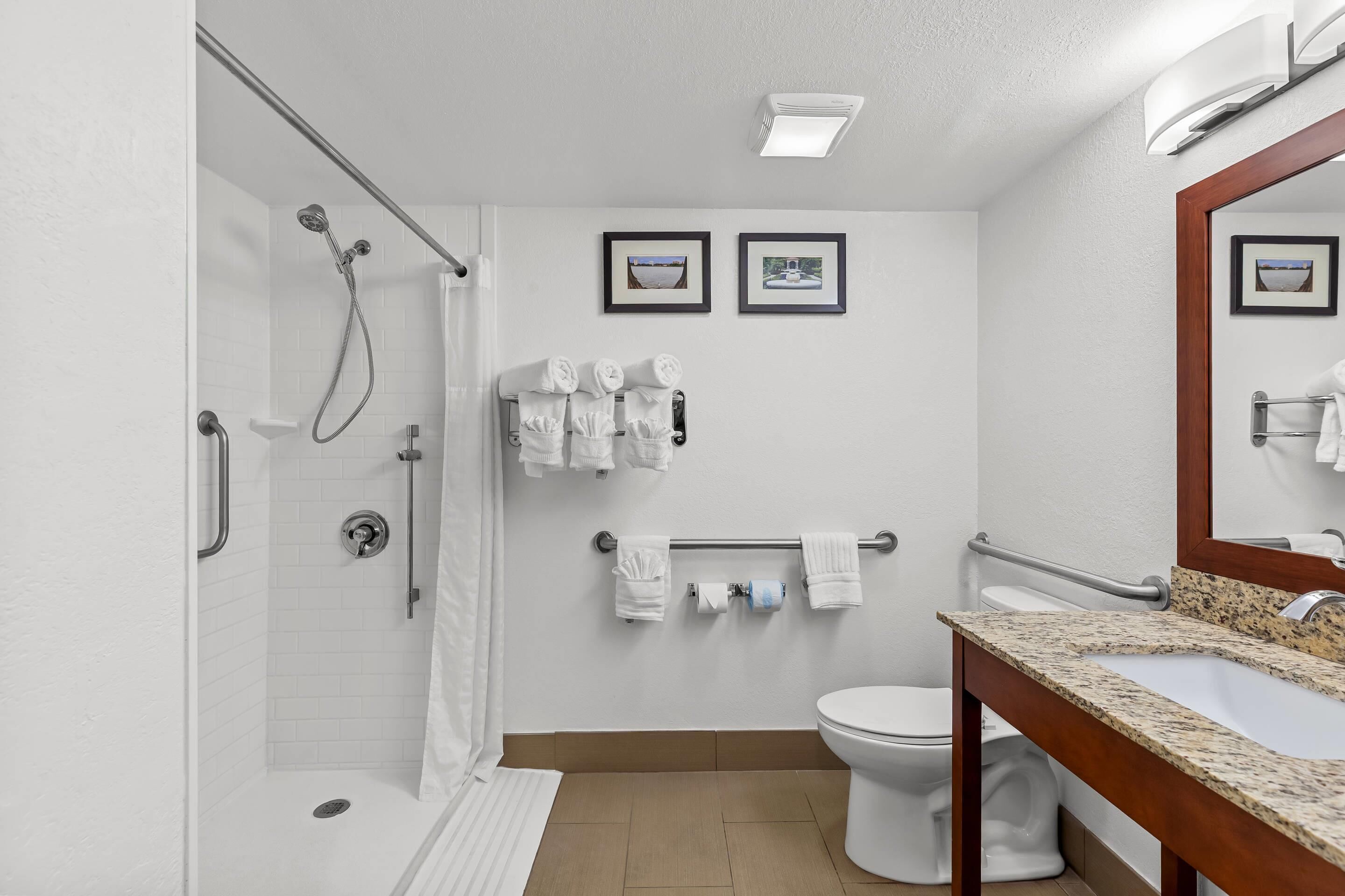 Un baño moderno con ducha, inodoro y lavamanos, equipado con barras de apoyo y un perchero para toallas.