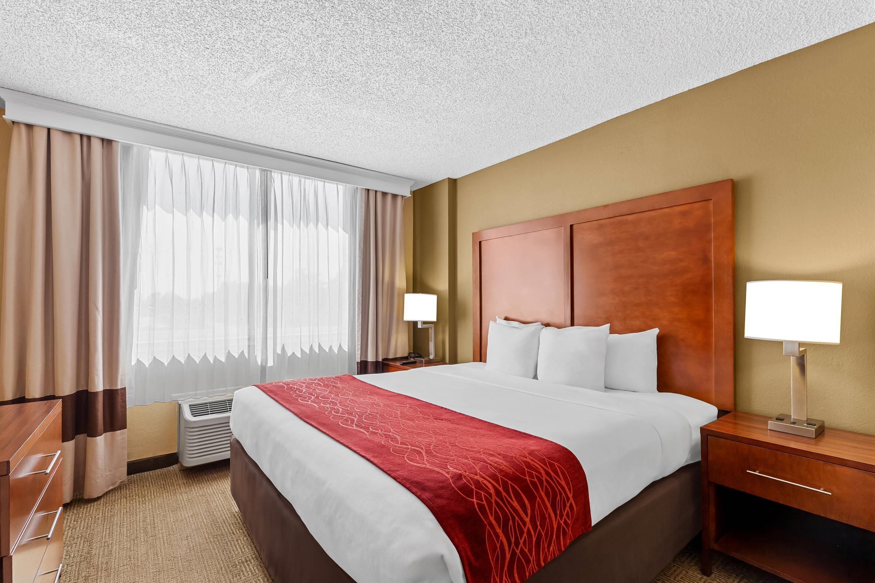Habitación de hotel con una cama grande, dos mesitas de noche con lámparas, cabecero de madera, ventana con cortinas y suelo alfombrado.