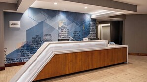 A modern reception desk with a wooden finish and a sloped design, situated in a hotel lobby with a geometric wall design and a sign indicating the hotel name 'Marriott Bonvoy'.