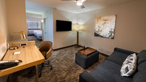 A hotel room with a desk, a sofa, a TV, a ceiling fan, a lamp, and a painting on the wall.