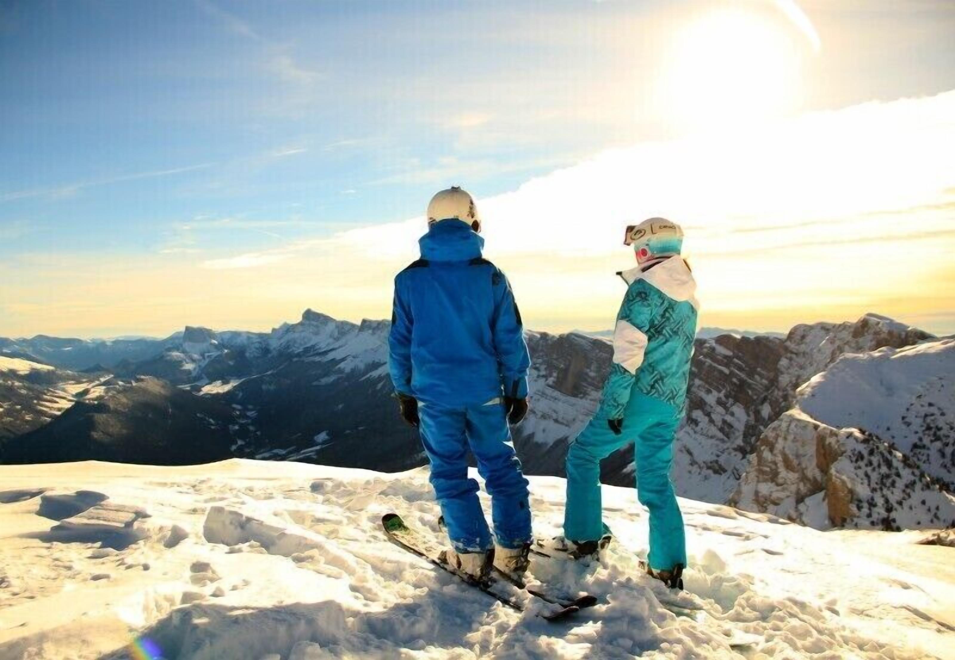 Two people standing on a snowy mountain peak with a view of distant mountains and a clear sky.