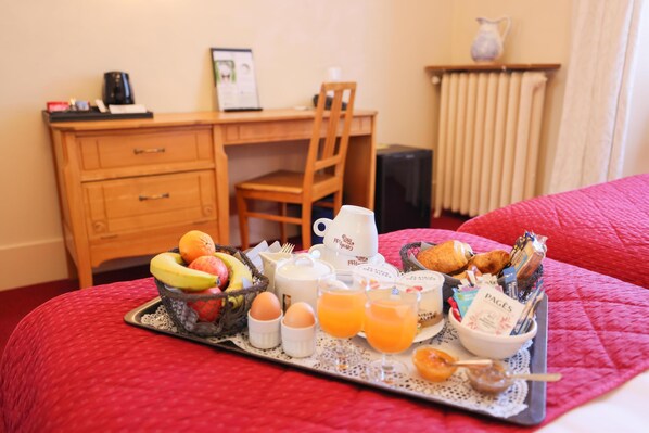 Bandeja de desayuno con frutas, huevos, jugo de naranja y pasteles sobre una cama con una colcha roja.