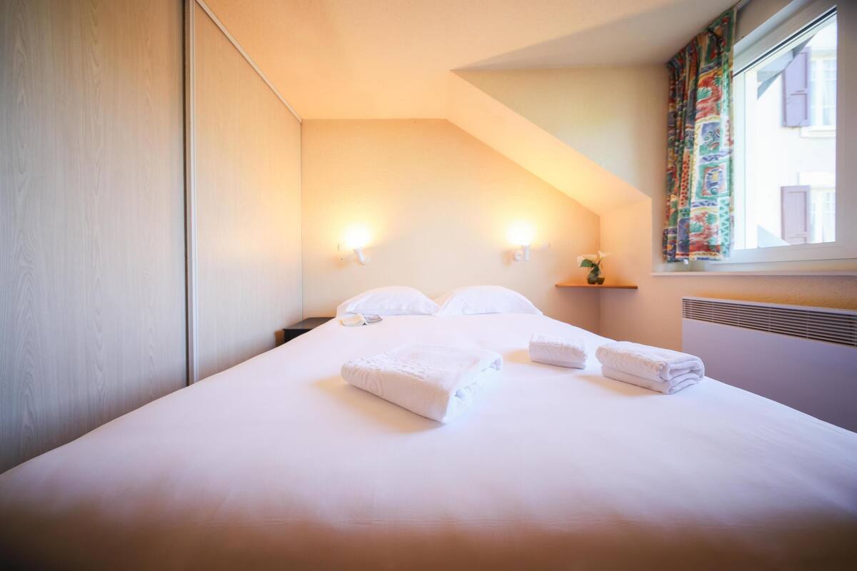 A hotel room with a neatly made bed, two wall-mounted lights, a window with curtains, and a small shelf with a plant.