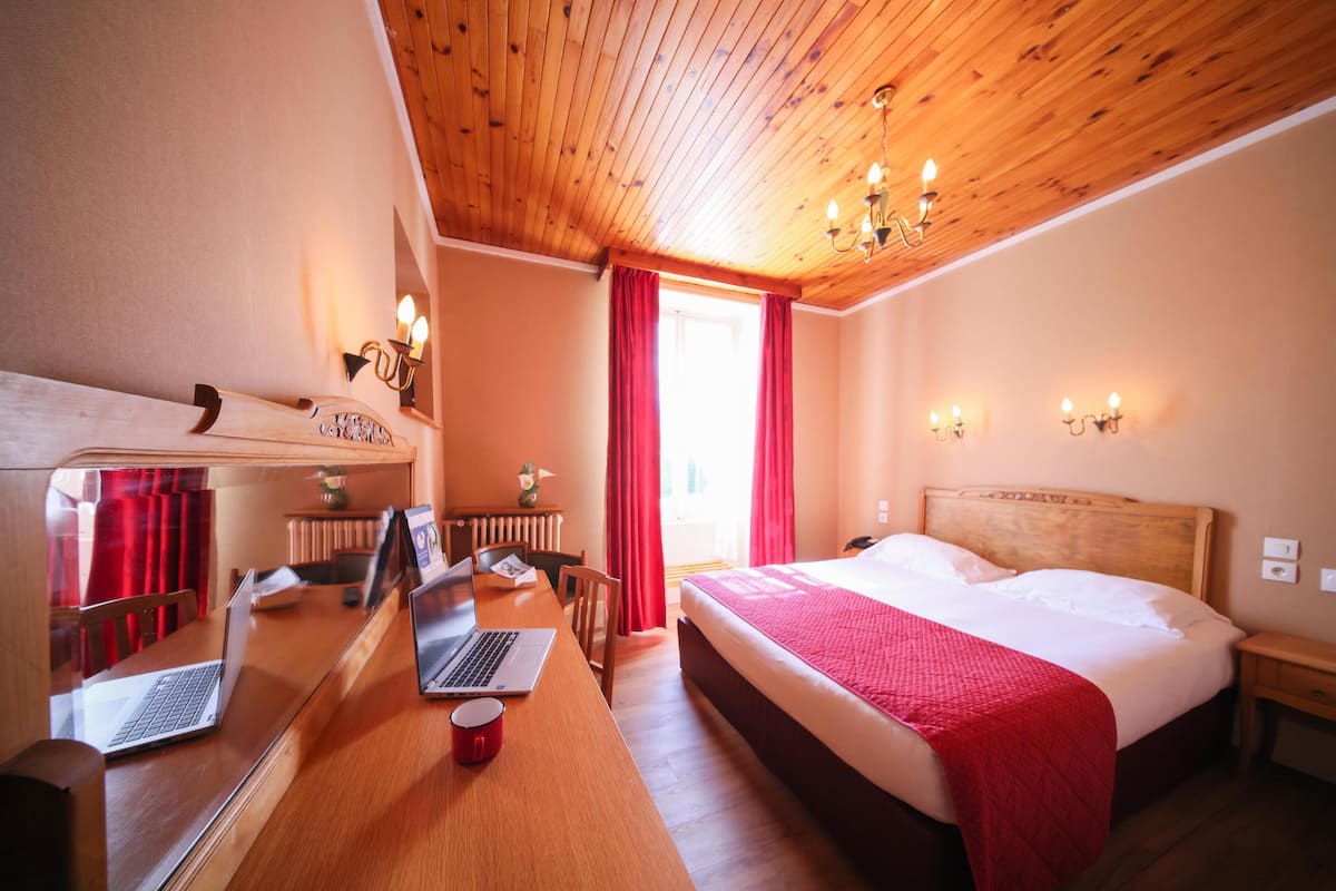 A hotel room with a wooden ceiling, a bed with a red blanket, a desk with a laptop, and a window with red curtains.