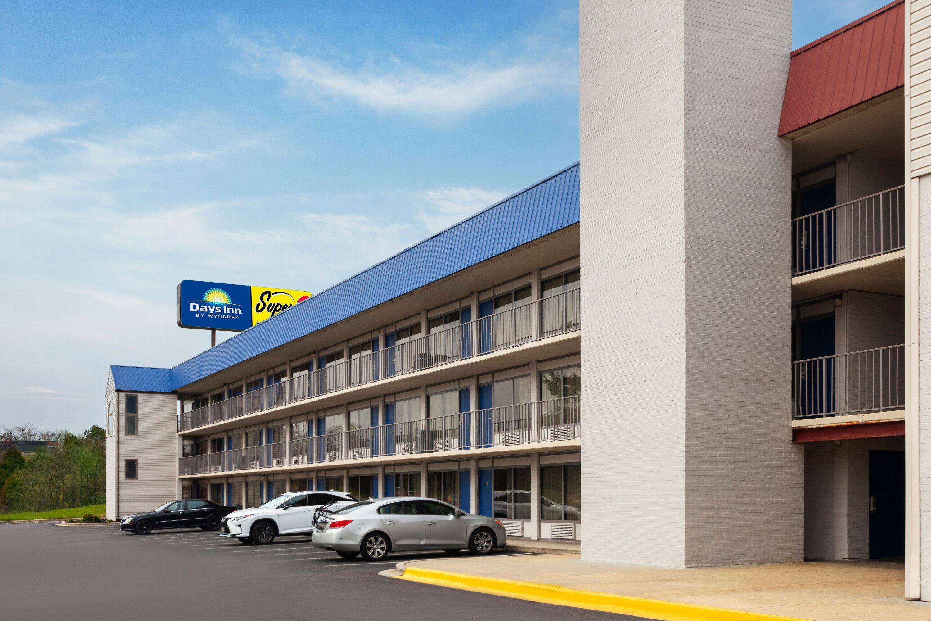 Een motel met een blauw dak en een bord met 'Super 8 by Wyndham'. Er zijn meerdere verdiepingen met balkons en een parkeerplaats met verschillende auto's.