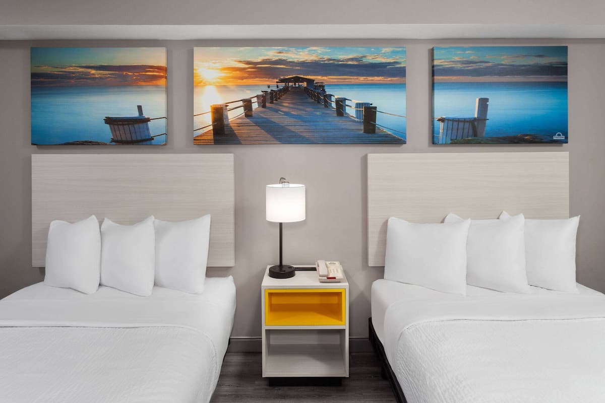 A hotel room with two beds, a nightstand with a lamp, and three paintings of a pier and sunset on the wall.