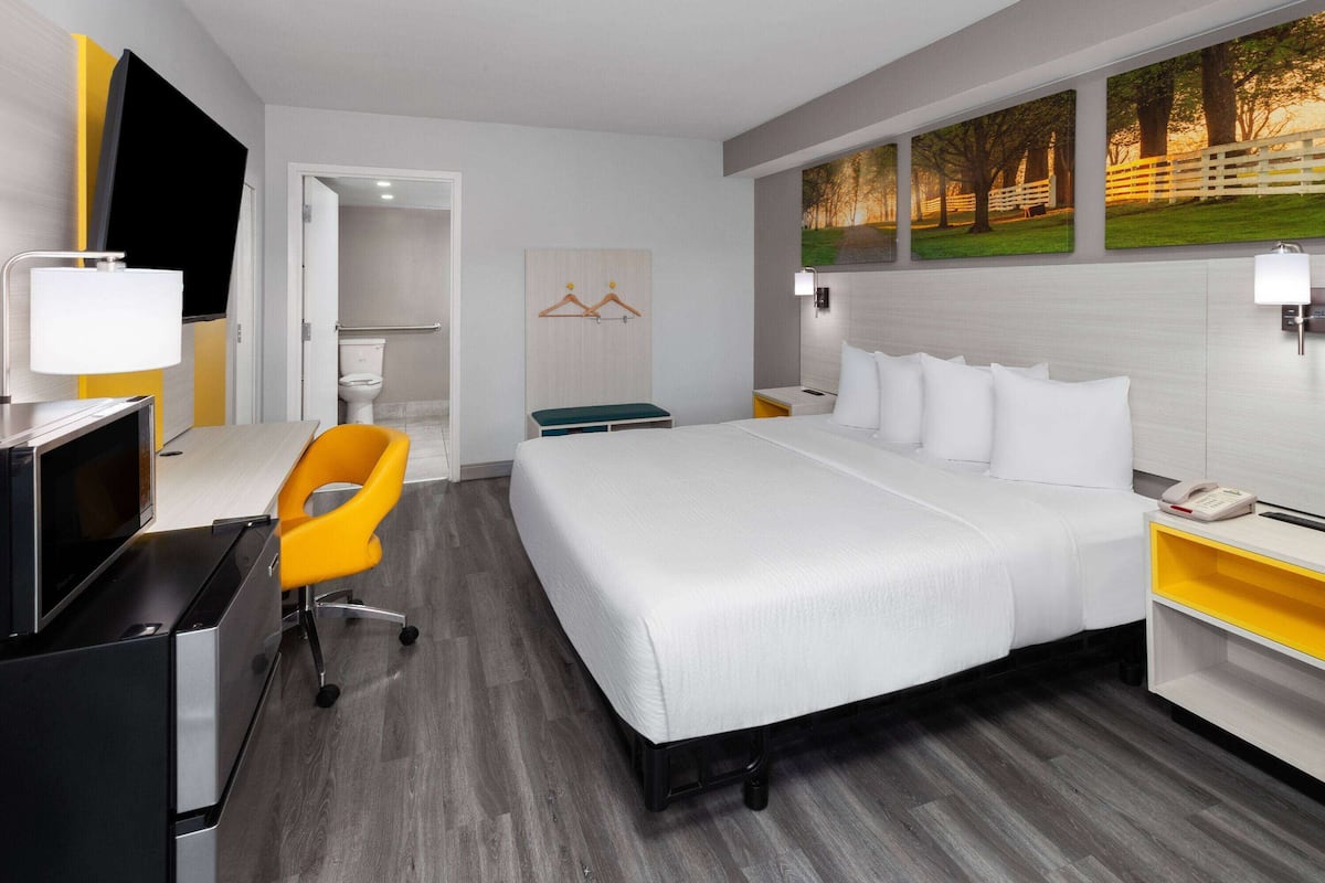 A hotel room with a bed, a desk with a computer, a chair, a bedside table with a phone, and artwork on the wall.