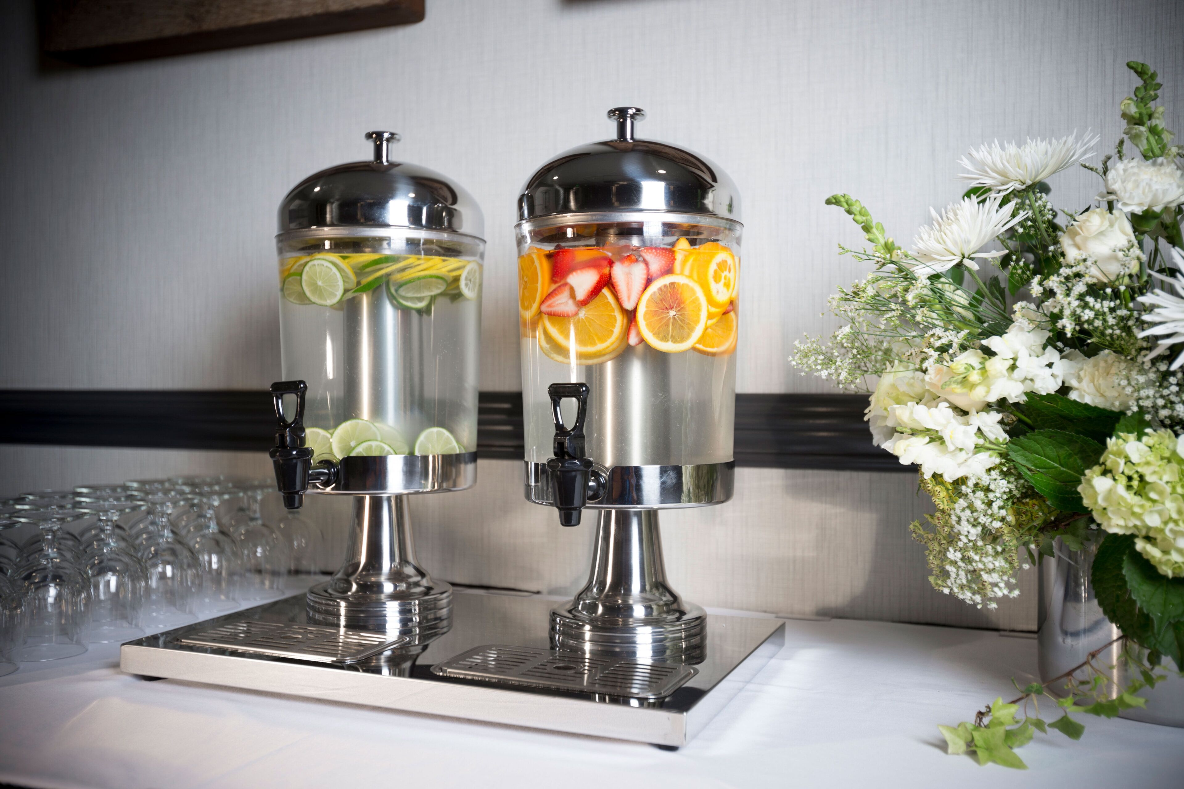 Two stainless steel beverage dispensers with fruit slices on top, a glass tray, and a floral arrangement.