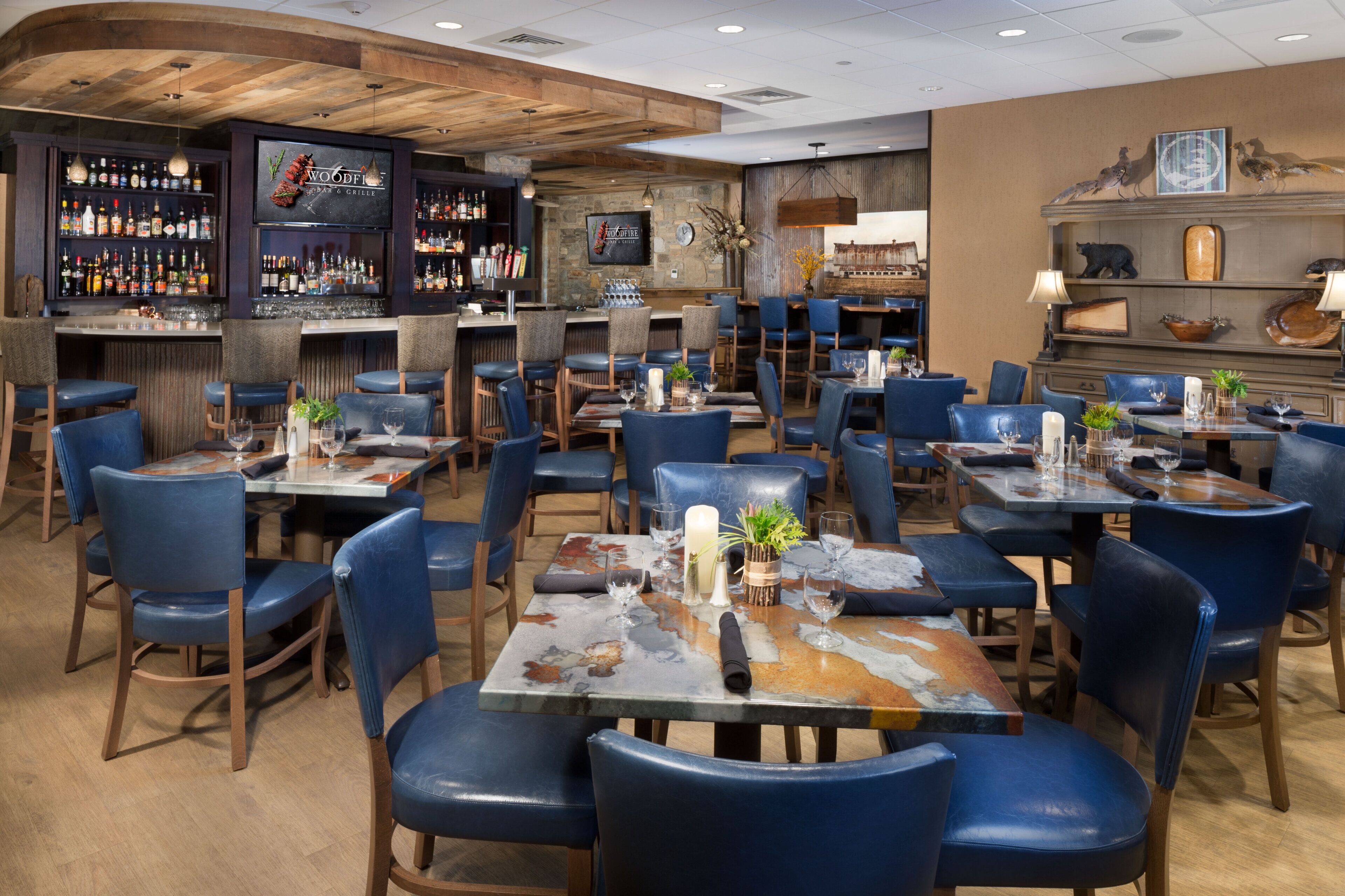 A restaurant interior with multiple tables and chairs, a bar area with a variety of bottles, and a rustic wooden ceiling.