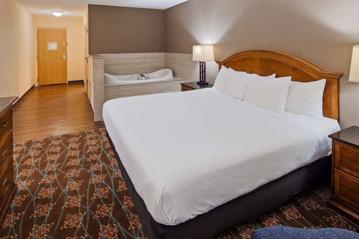 A hotel room with a large bed, a separate seating area with a chair, a small table, a nightstand, a lamp, and a bathroom with a bathtub.