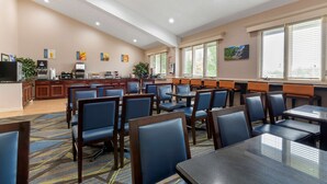 A hotel dining area with multiple tables and chairs, a kitchen area, and large windows with blinds. - Best Western University Inn (Ithaca)