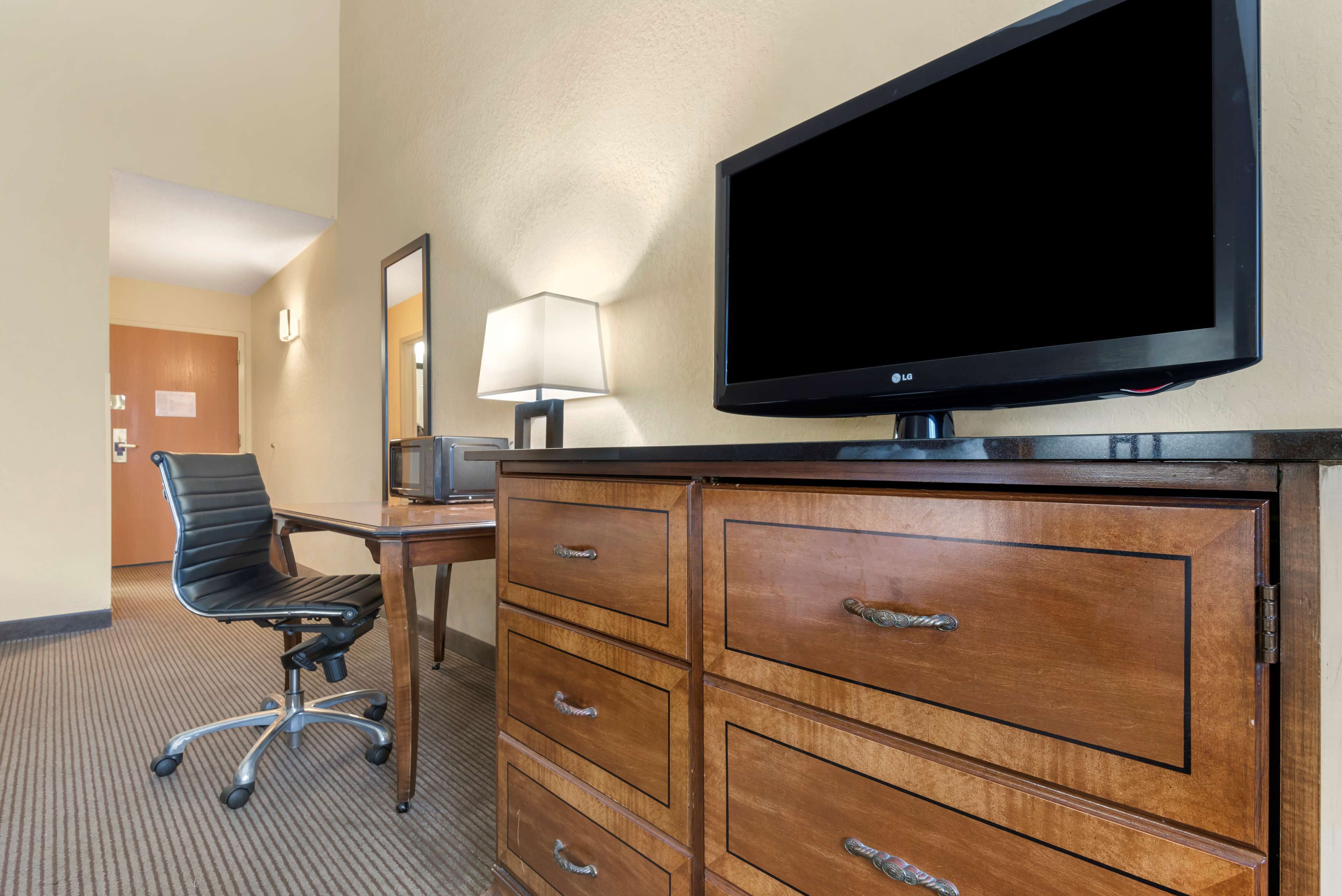 A hotel room with a flat-screen TV, a wooden dresser with drawers, a black office chair, and a desk with a lamp.