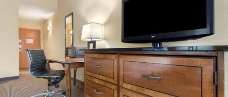 A hotel room with a flat-screen TV, a wooden dresser with drawers, a black office chair, and a desk with a lamp.