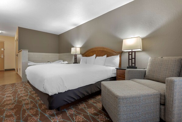 Une chambre d’hôtel avec un grand lit, deux lampes de chevet, un fauteuil gris et un tapis à motifs.