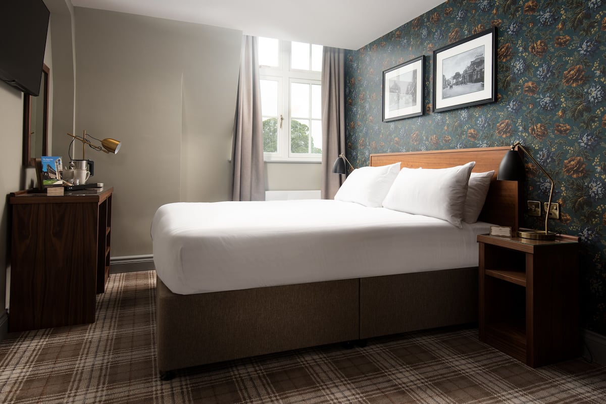A neatly made bed in a room with a plaid carpet, a wooden nightstand, a lamp, and two framed pictures on the wall.