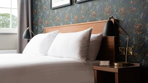 A neatly made bed with white linens and two pillows, a wooden nightstand with a lamp, and a wall with floral wallpaper. - The Ripon Inn - The Inn Collection Group (Ripon)