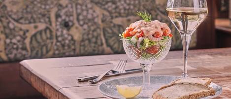 Una mesa con un bol de ensalada de vidrio, una copa de vino blanco y una rebanada de pan en un plato.