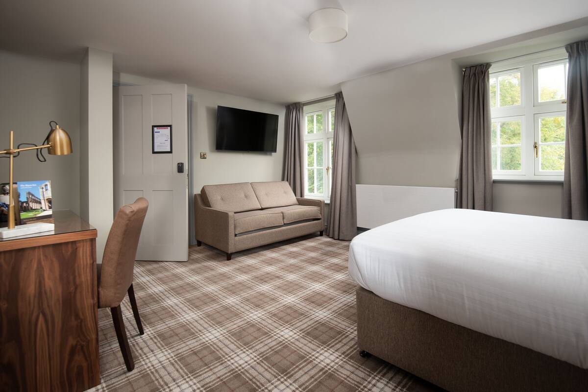 A hotel room with a bed, a sofa, a desk with a lamp, a television, and a window with curtains.