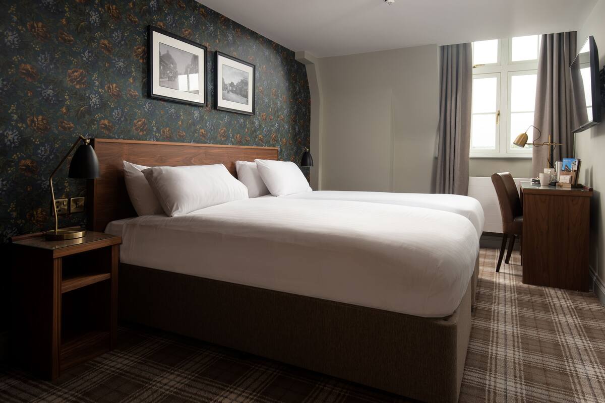 A hotel room with a large bed, a desk, a chair, a lamp, and two framed pictures on the wall.