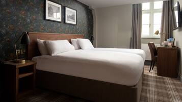 A hotel room with a large bed, a desk, a chair, a lamp, and two framed pictures on the wall.
