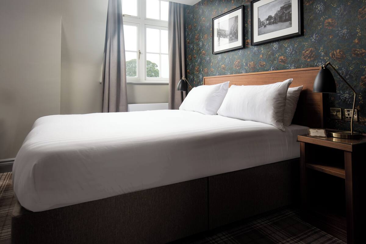 A neatly made bed in a hotel room with a wooden headboard, a nightstand with a lamp, and two framed pictures on the wall.