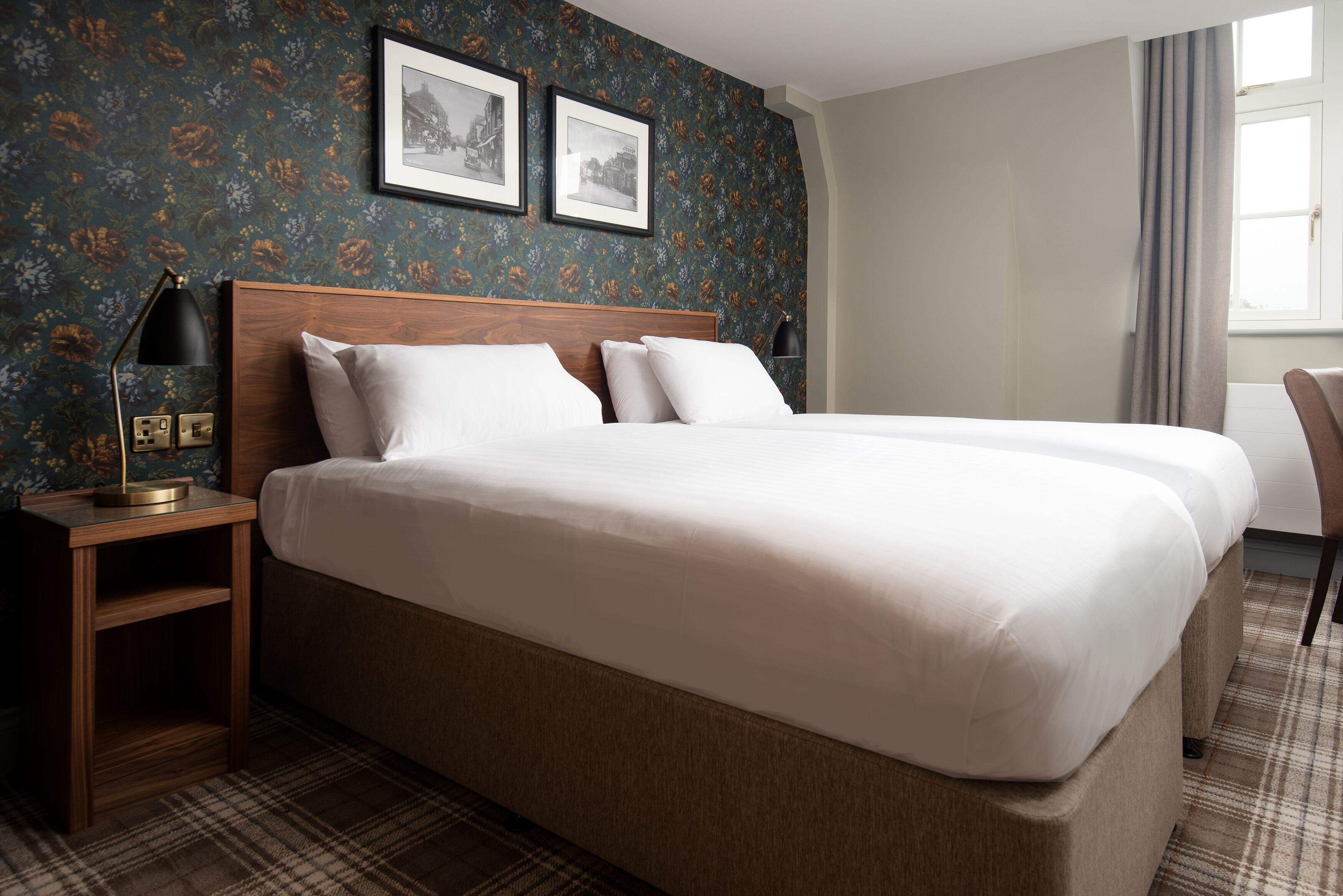 A double bed with a wooden headboard, a nightstand with a lamp, and a plaid-patterned rug.