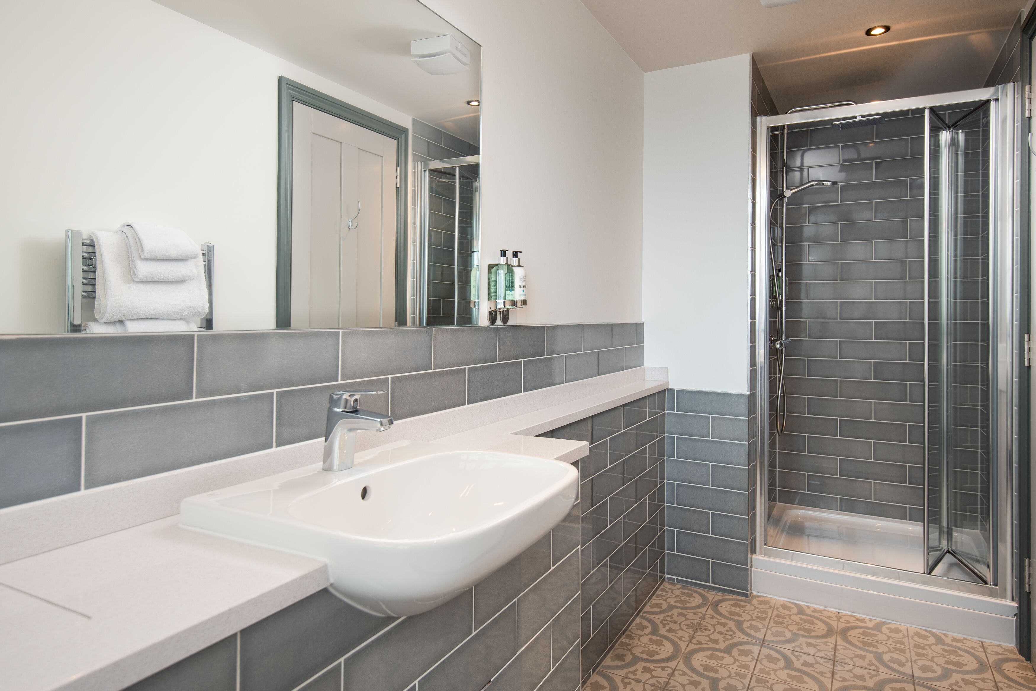 A modern bathroom with a white sink, grey tiles, a glass shower enclosure, and a mirror with a towel rack.
