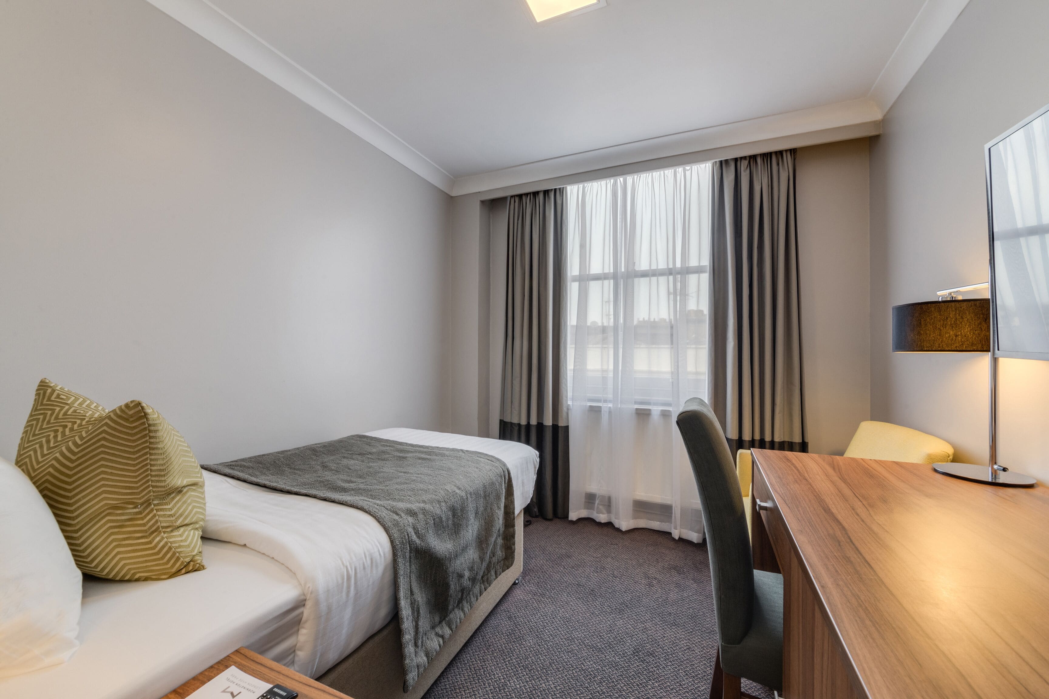 A hotel room with a bed, a desk, a chair, a lamp, and a window with curtains.